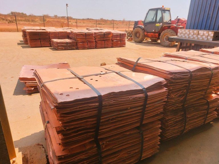 Stacks of bundled brown metal sheets or plates on a construction yard, bound with black straps, with a red forklift in the background.