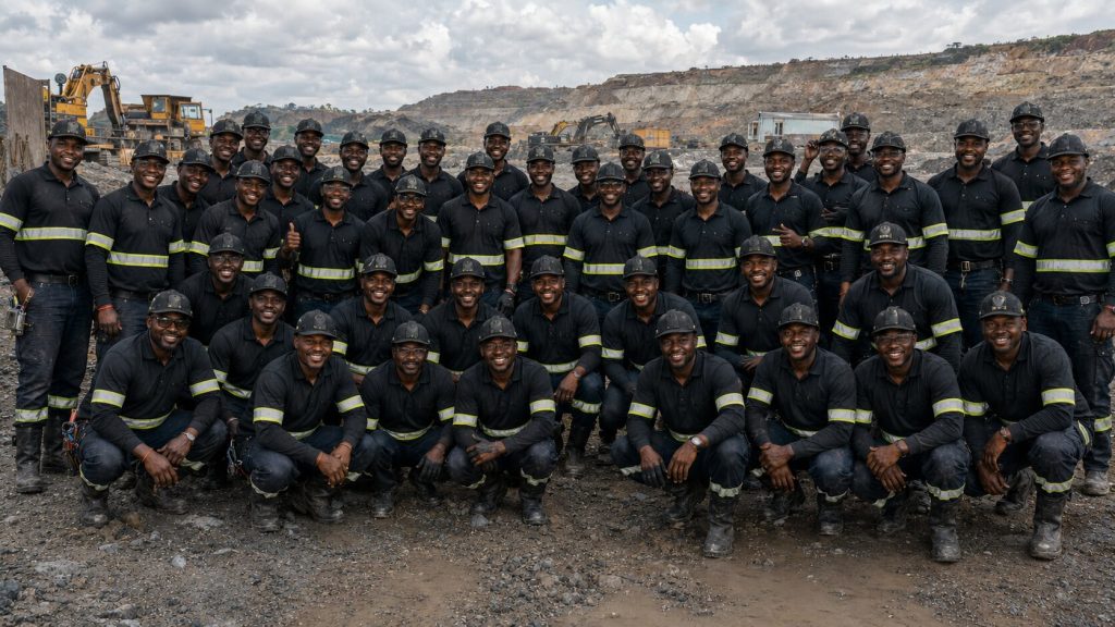 Group of firefighters in black turnout gear with reflective stripes posing for a team photo at a rocky quarry site with heavy machinery in the background.