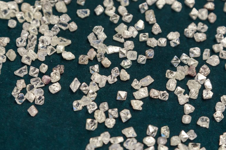 Many small translucent crystals scattered on a dark teal surface, resembling coarse salt or sugar crystals.